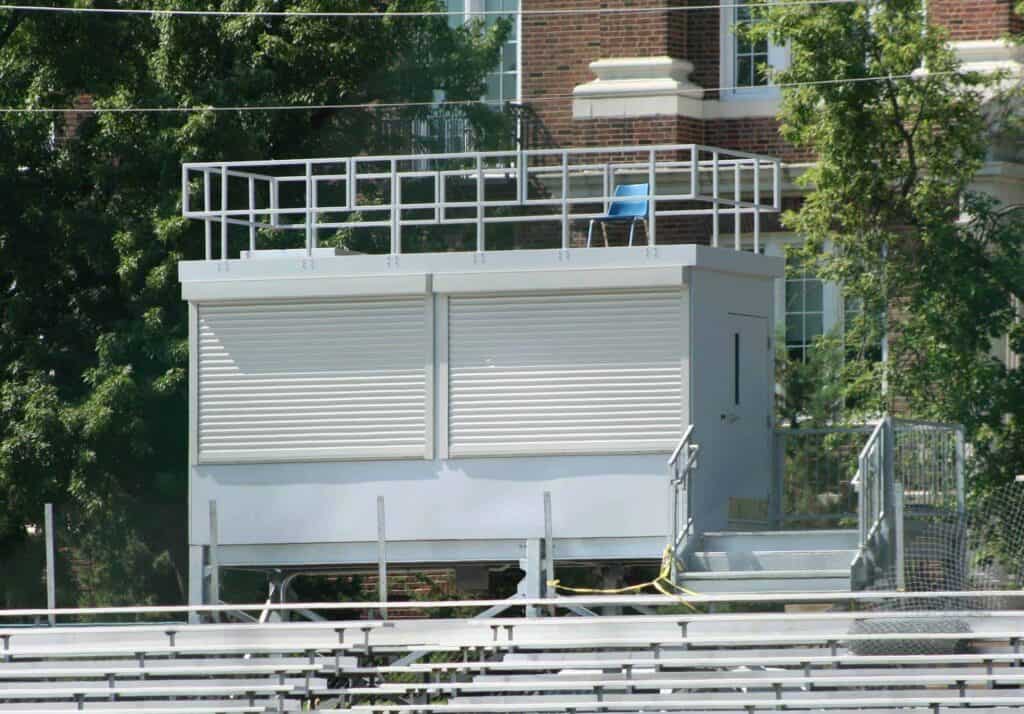 A press box at a sports field, dedicated to sports journalism, is situated above empty metal bleachers. It features a white exterior with a flat roof and large windows covered by shutters. Trees and