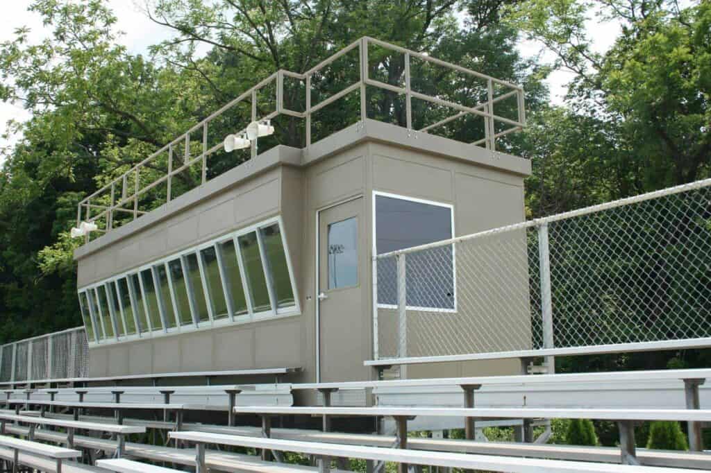 A small beige sports media box with large windows sitting above bleachers next to dense trees.