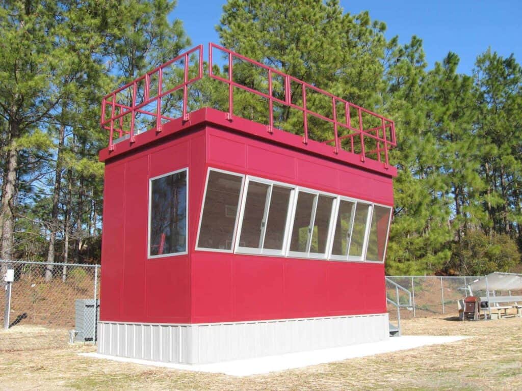 A red, rectangular outdoor press box with large windows and a flat roof, equipped with a railing, situated in a grassy area with clear skies and sparse trees in the background.