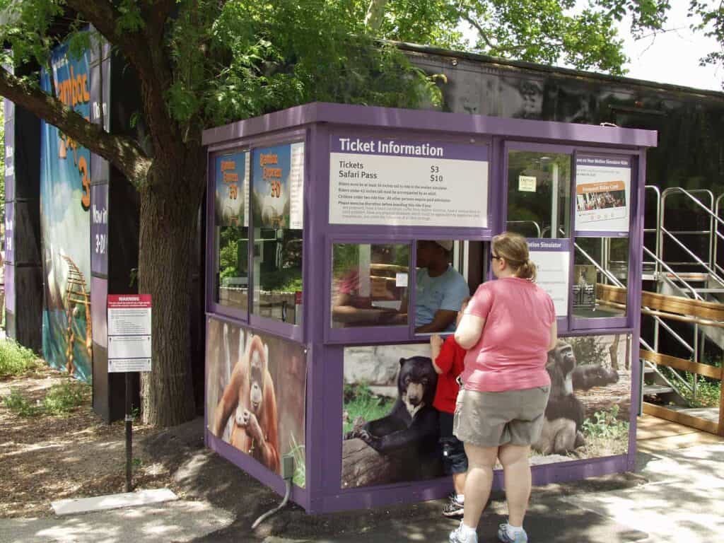 A woman stands at a zoo ticket booth, talking to the cashier inside. The booth is adorned with animal images and displays prices for tickets and safaris. Trees surround the area.