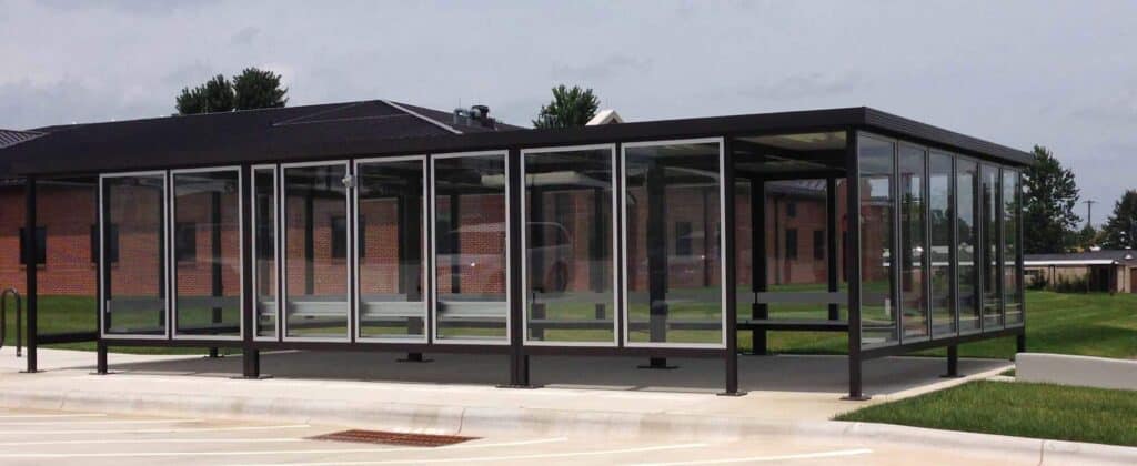 Smoking Shelter 1 A modern glass-enclosed walkway connecting two buildings on a campus, set against a cloudy sky with smoking shelters and a lawn visible in the background.