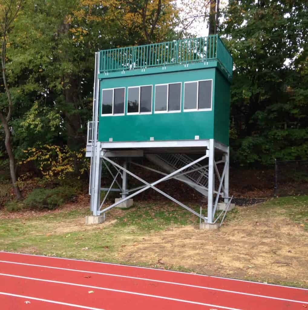 Elevated press box for sports journalism with windows, situated beside a red running track, surrounded by trees in an outdoor sports area.