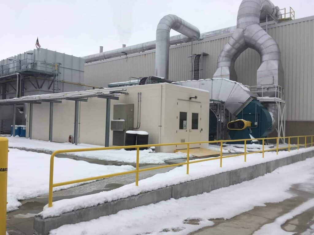 An industrial facility in winter with snow-covered ground. The building has large pipes and ducts, a blue vent, and is surrounded by safety yellow railings and equipment enclosures.