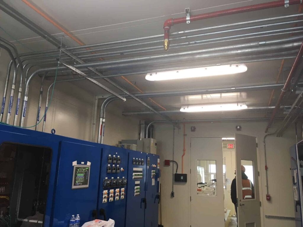 A well-organized industrial workspace with blue machinery, neat electrical wiring, and outdoor equipment enclosures overhead, bright overhead lights, and a person in the distance at the exit door.