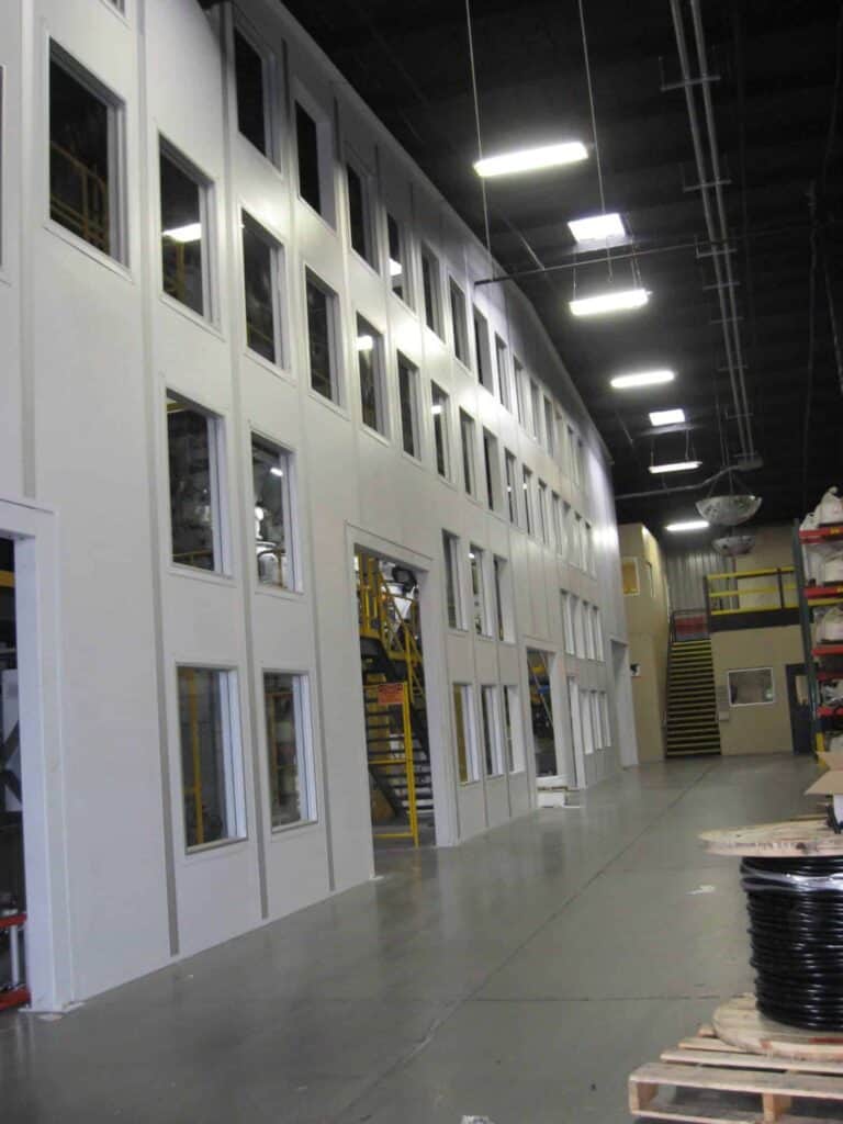 Tall Modular Wall With Windows 10 Interior of an industrial facility showing a row of windows with a view inside various rooms, a yellow staircase, and materials arranged in the foreground, bordered by tall walls.