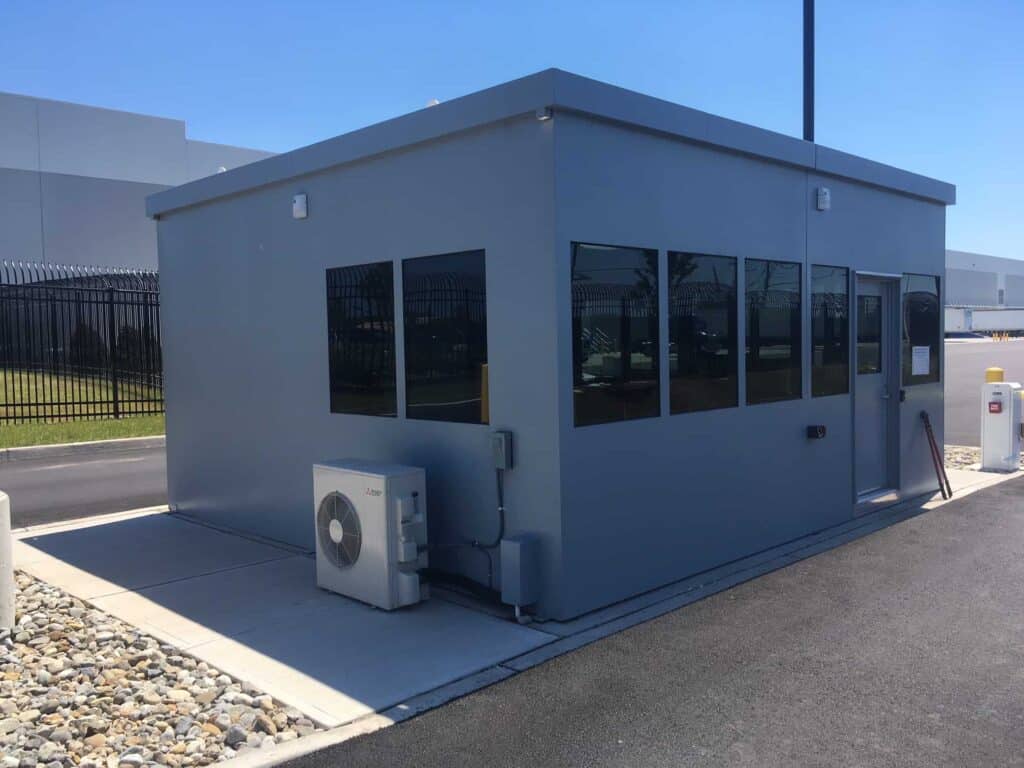 Guard/Security Booth 47 A small, modern gray security kiosk with multiple windows, installed at an industrial site. The kiosk is equipped with an air conditioning unit on its side, situated on a concrete pad with rocks around