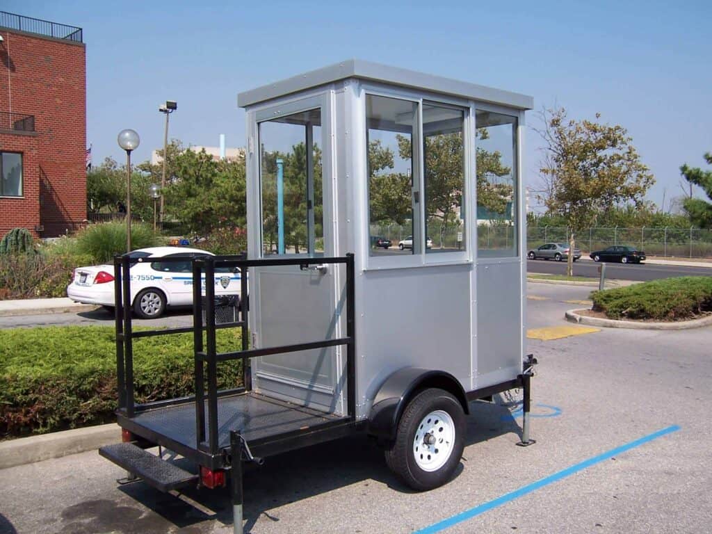 Guard/Security Booth 12 A small, mobile security booth with a window and a door on a single-axle trailer, parked in an outdoor area with trees and buildings in the background.
