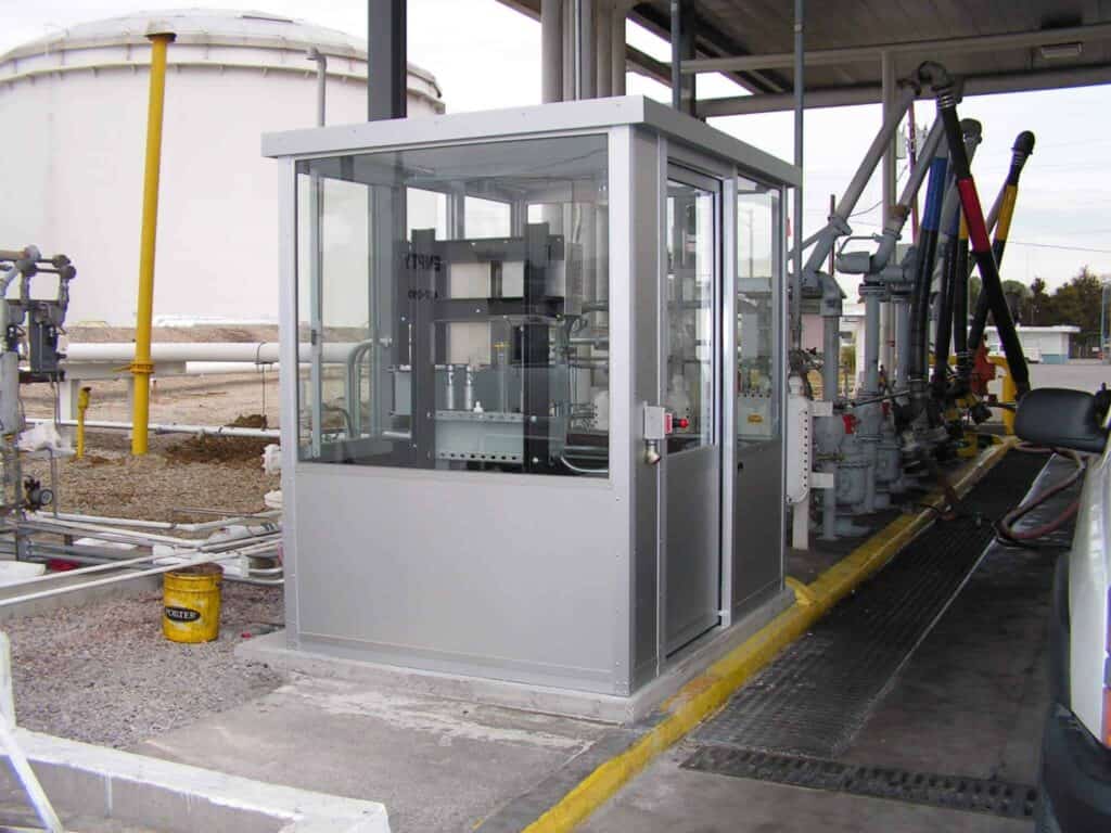 A small, enclosed industrial workstation with automated machinery, surrounded by various outdoor equipment enclosures and large storage tanks in an outdoor setting.