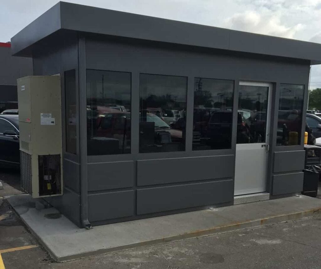 Guard/Security Booth 19 A small, modern gray building with large glass windows and a security booth attached on the left side, located in a parking lot with cars reflected in the glass.