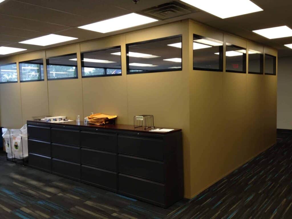 A modern office with a large black storage cabinet, a sectioned beige cubicle with modular partitions and glass on the top half, and a clean environment illuminated by overhead lights.