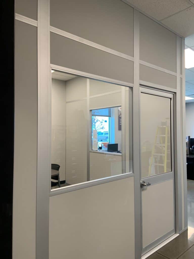 An office with glass wall partitions and doors, showing a computer monitor inside. Reflections of the exterior, including a parked car, are visible on the glass.