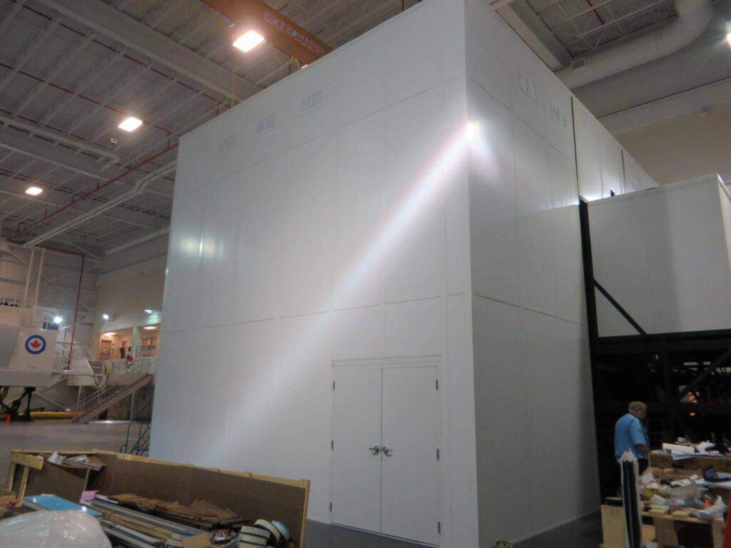 Large industrial white structure with a beam of light shining on it, located in a spacious warehouse with construction materials scattered around and a person in the background. This structure is one of the indoor equipment enclosures