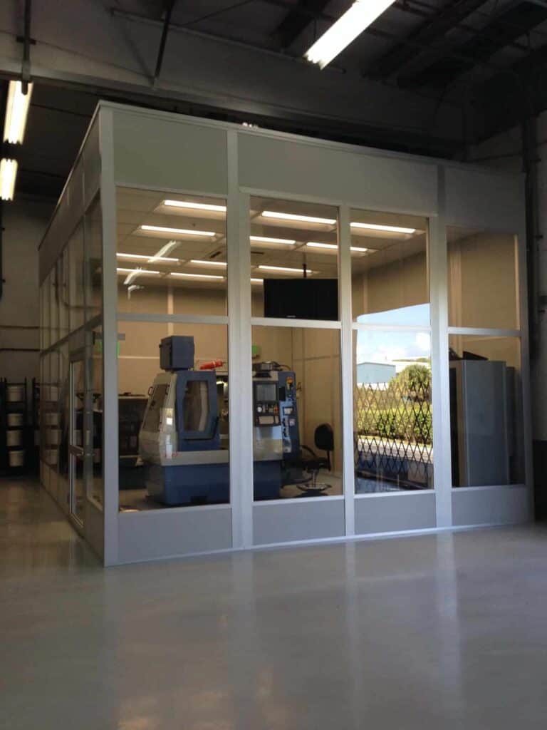 A glass-walled office inside an industrial workshop, showcasing a clear view of machinery and indoor equipment enclosures, illuminated by natural light.