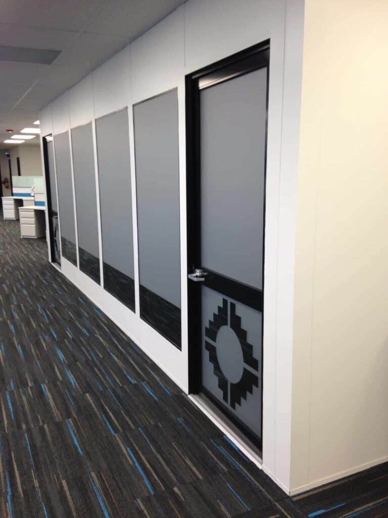 A modern office space with a series of large frosted glass doors along a white wall, featuring modular office partitions and a black and gray gear logo on one door. The floor is carpeted in blue