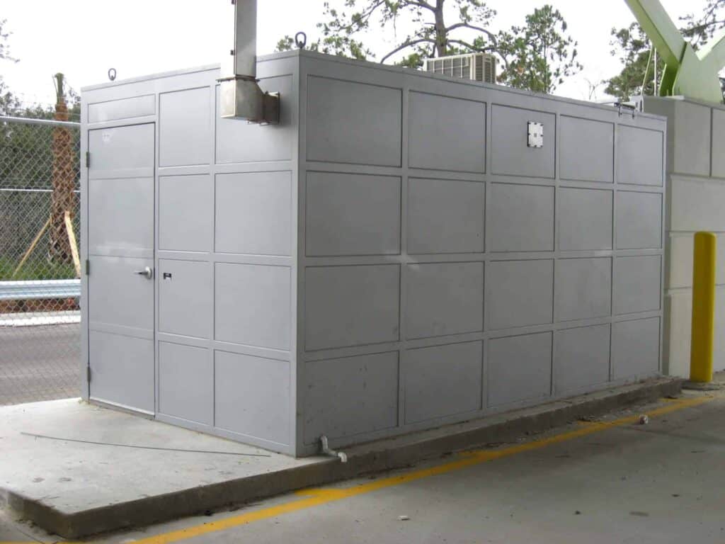 Large gray outdoor equipment enclosure with multiple panels, situated in a paved area near a road, with trees in the background and a yellow pole on the left.