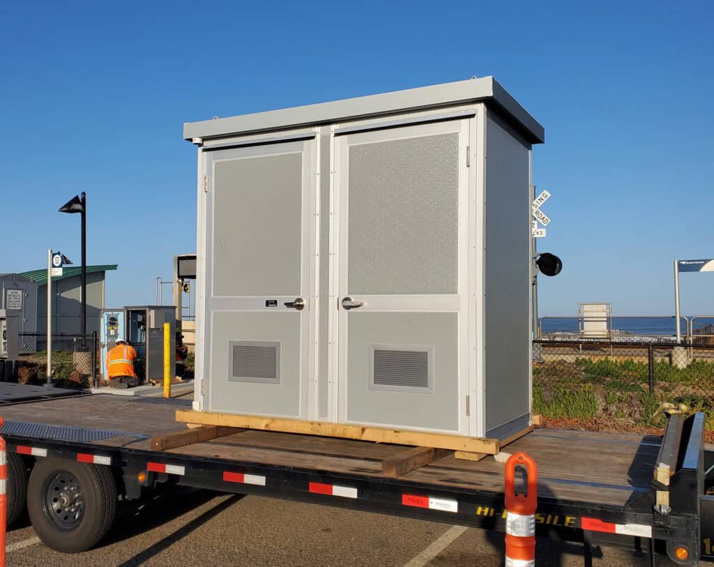 A small gray utility shed with double doors and vent panels, mounted on a trailer in a sunny outdoor setting with ocean views in the background is an ideal example of outdoor equipment enclosures.