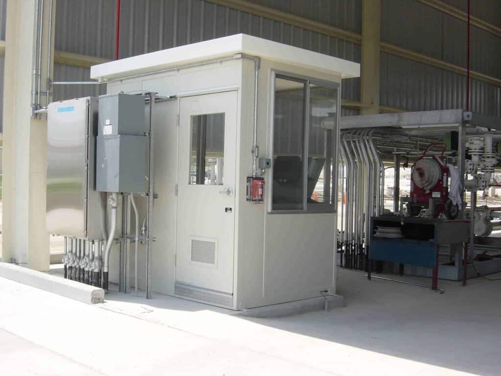 A small industrial control room with a large electrical equipment enclosure next to it, located outside under a partial overhang, with industrial pipes and machinery nearby.