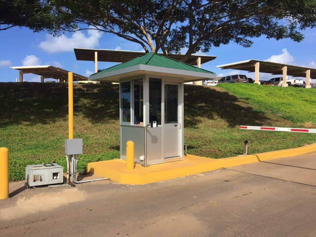 Guard/Security Booth 28 A bright sunny day at a parking lot entrance featuring a small safety booth with a raised barrier arm, surrounded by greenery and parked cars in the background.