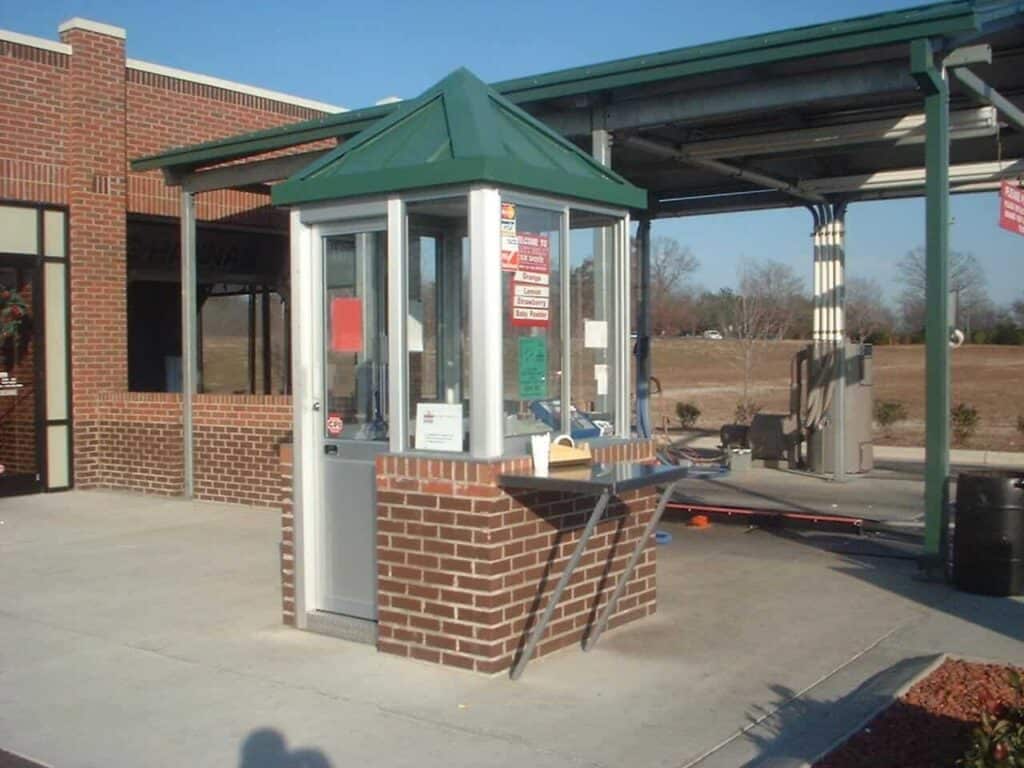 A small brick-built ticket booth with a green metal roof, featuring a window and an open door. The booth is located next to a sheltered area with a metal framework.