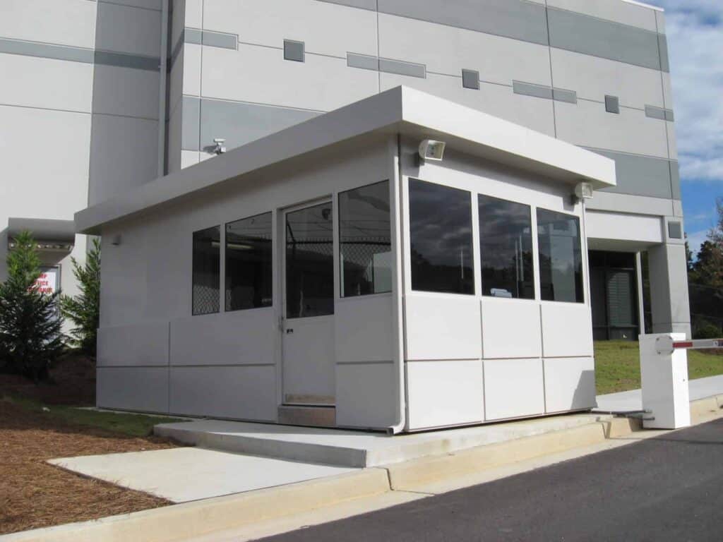 Guard/Security Booth 16 A small, modern security booth positioned outside a large building with glass-paneled sides and a metal door, under a clear sky. The booth is equipped with outdoor lighting and is adjacent to a driveway for