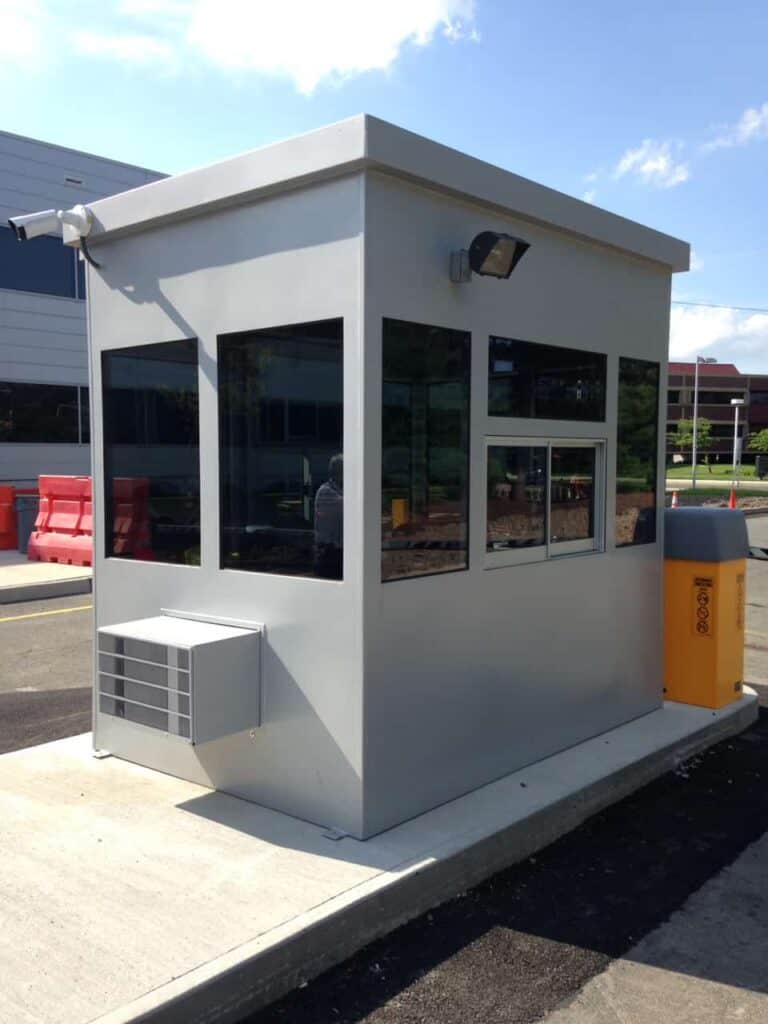 Guard/Security Booth 7 A small access control booth with large windows, stationed at a parking lot entrance, equipped with an air conditioning unit and an adjacent yellow barrier arm.