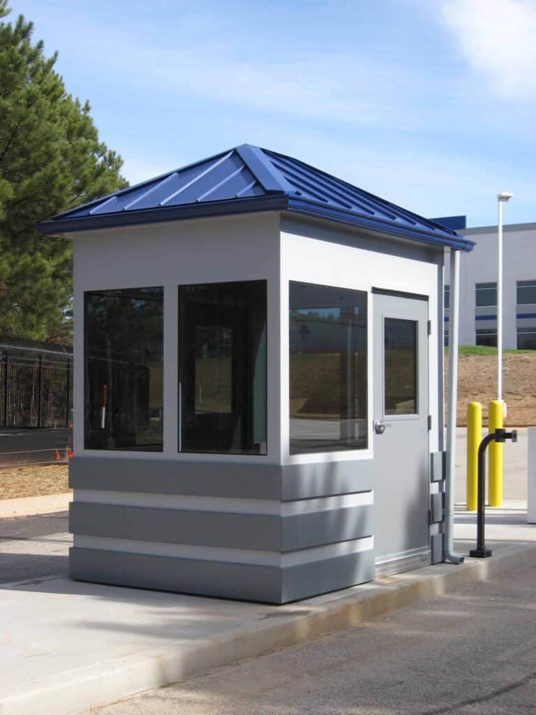 Guard/Security Booth 24 A small, modern security booth with large windows and a blue roof, situated on a concrete surface near a paved road and a fenced area.