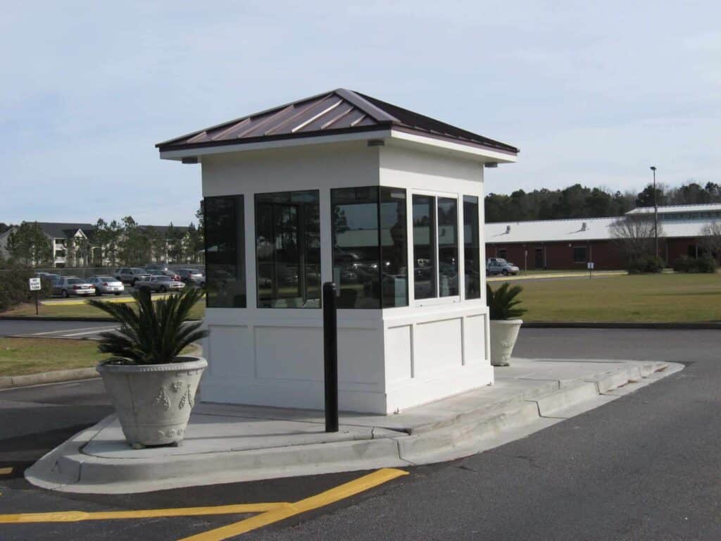Guard/Security Booth 20 A small, white security booth with a gray roof, surrounded by glass windows, situated in a parking lot with potted plants on either side.
