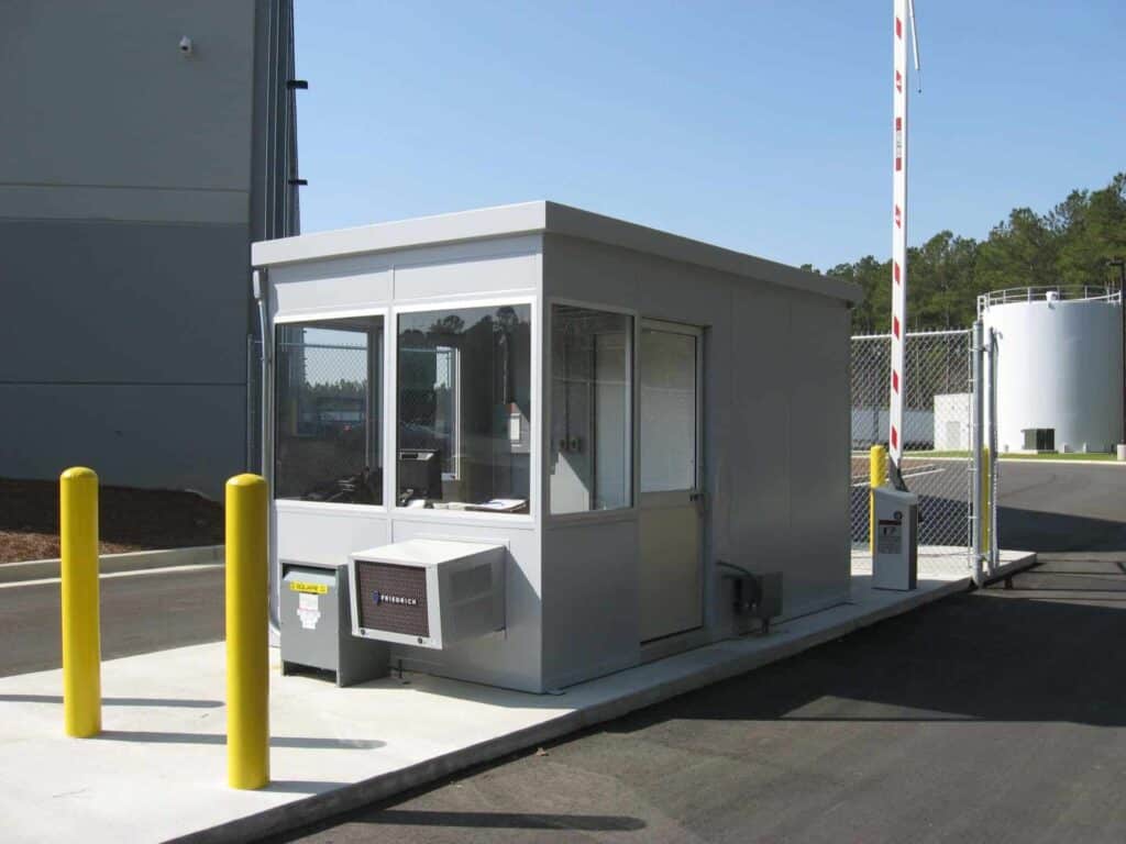 Guard/Security Booth 10 A small, modern portable security booth with large windows, located at an industrial facility entrance, flanked by yellow safety bollards and set against a clear blue sky.