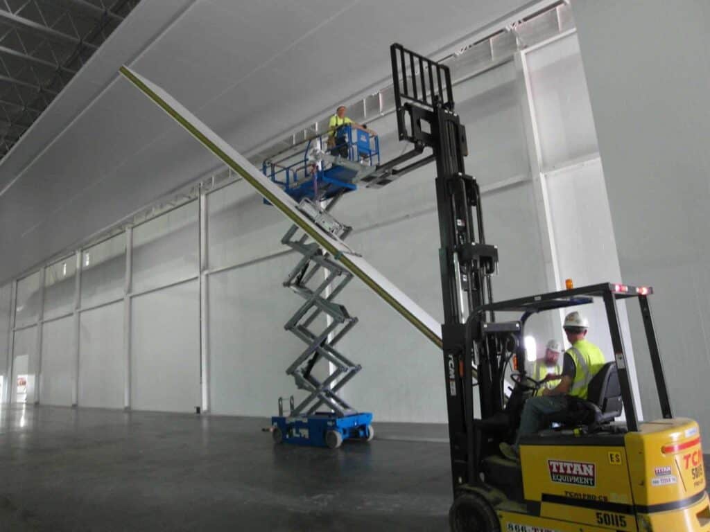 Tall Modular Wall Being Constructed Worker on a scissor lift installs lighting fixtures in a large industrial building with tall walls while another worker on a forklift assists by holding materials.