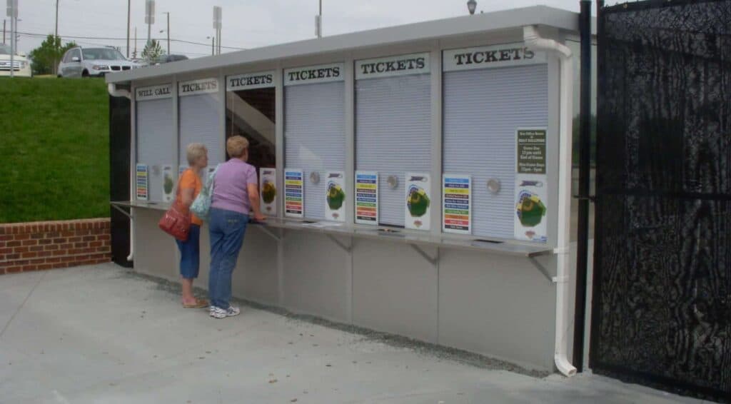 Two women standing at an outdoor ticket booth looking at event listings and prices posted under the window. The booth is gray with signs that read 